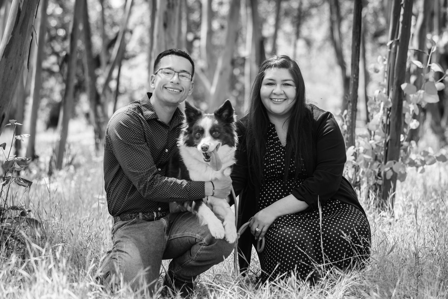 Black and white photo of a couple kneeling in a forest field with their border collie dog between them.