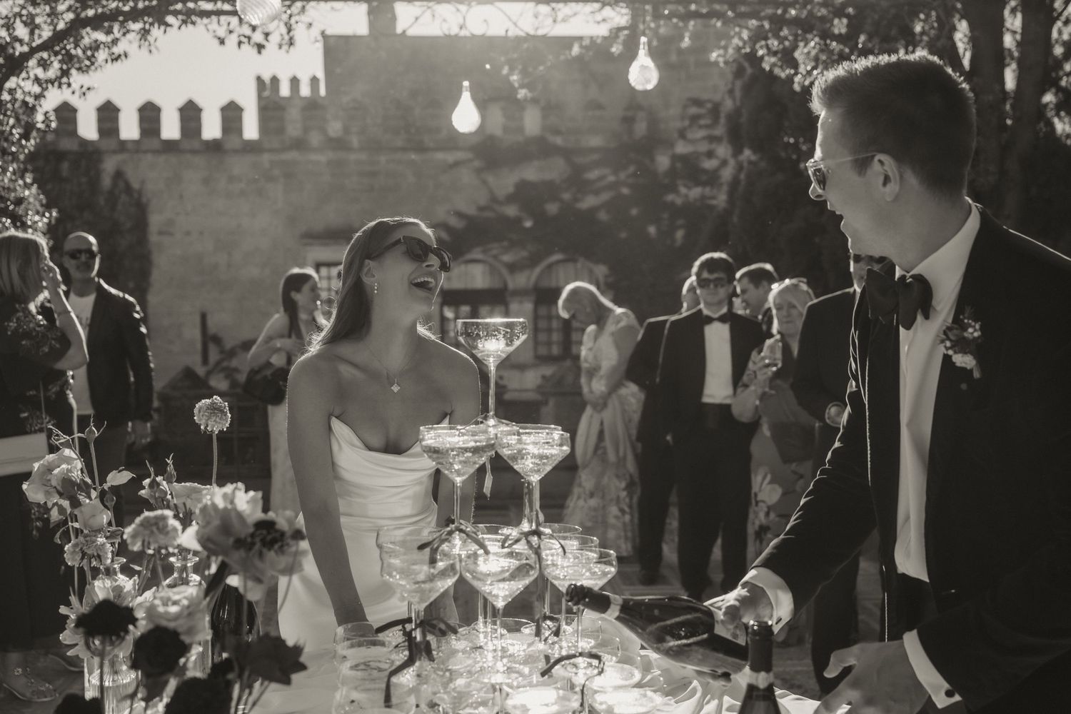 Black and white wedding photo of bride and groom smiling beside champagne tower at elegant outdoor reception.
