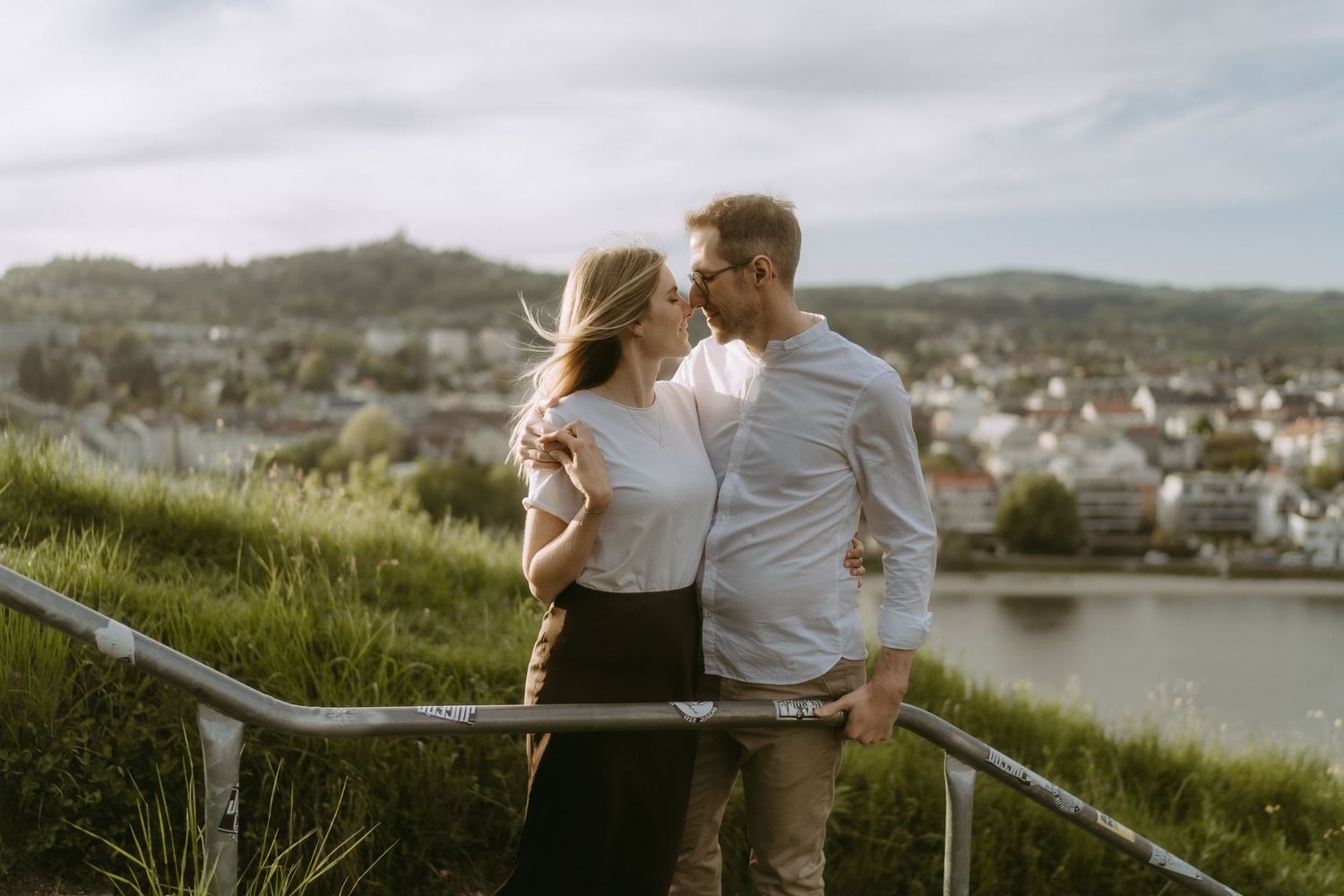 Romantic couple sharing a tender moment on a scenic overlook with a European town and river in the background.