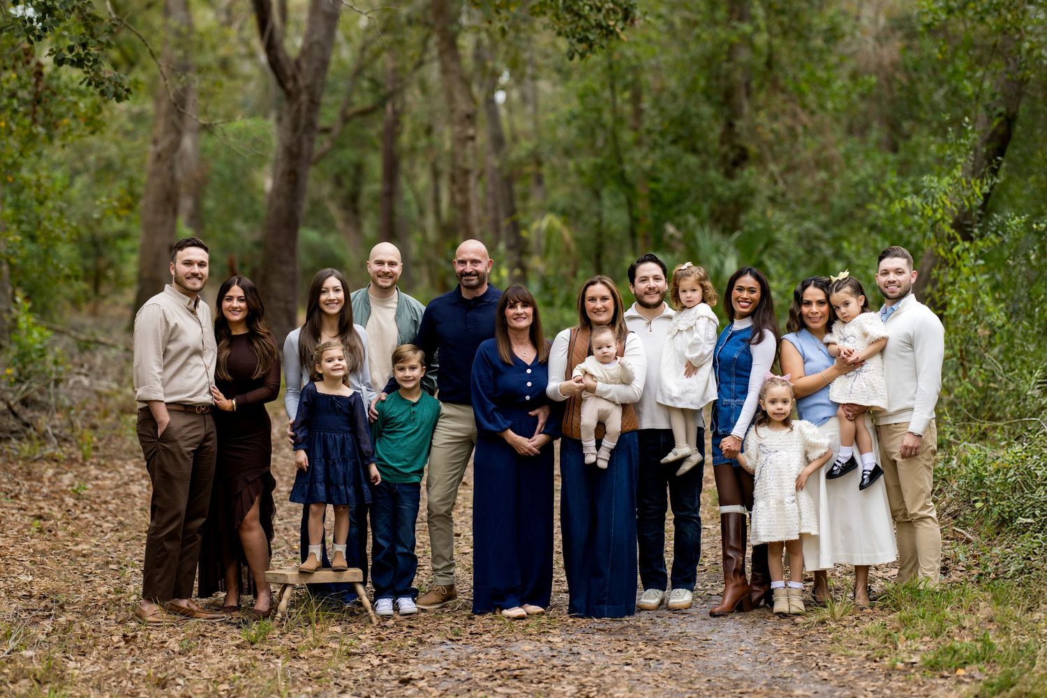 Large family group portrait outdoors on a wooded path, dressed in coordinated navy and neutral tones.
