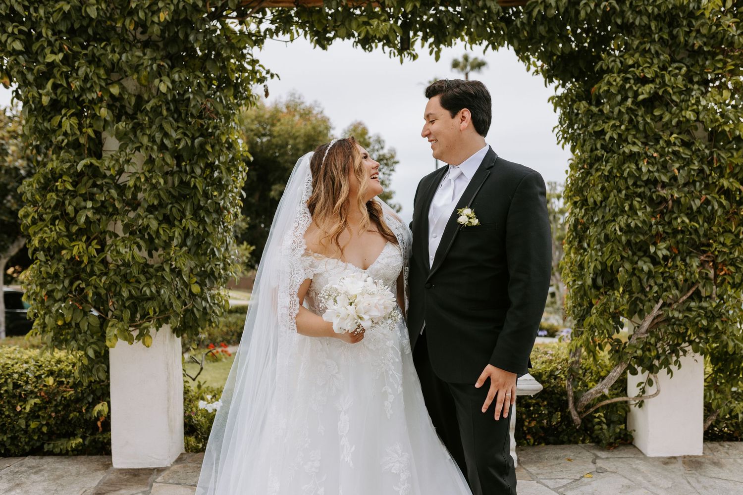 Bride and groom share a romantic moment under a greenery arch, bride in lace gown with veil holding white bouquet.