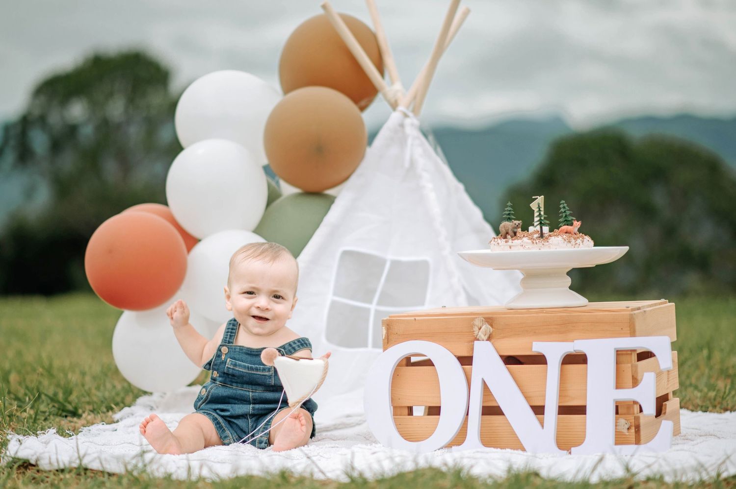 First birthday setup outdoors with white teepee tent, brown and white balloons, and ONE text decoration on white blanket.