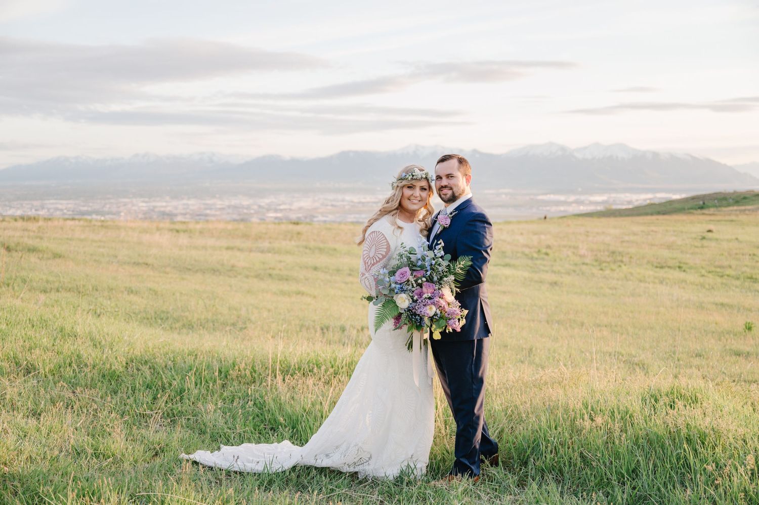 Scenic mountain wedding portrait with couple standing in grassy field at sunset.