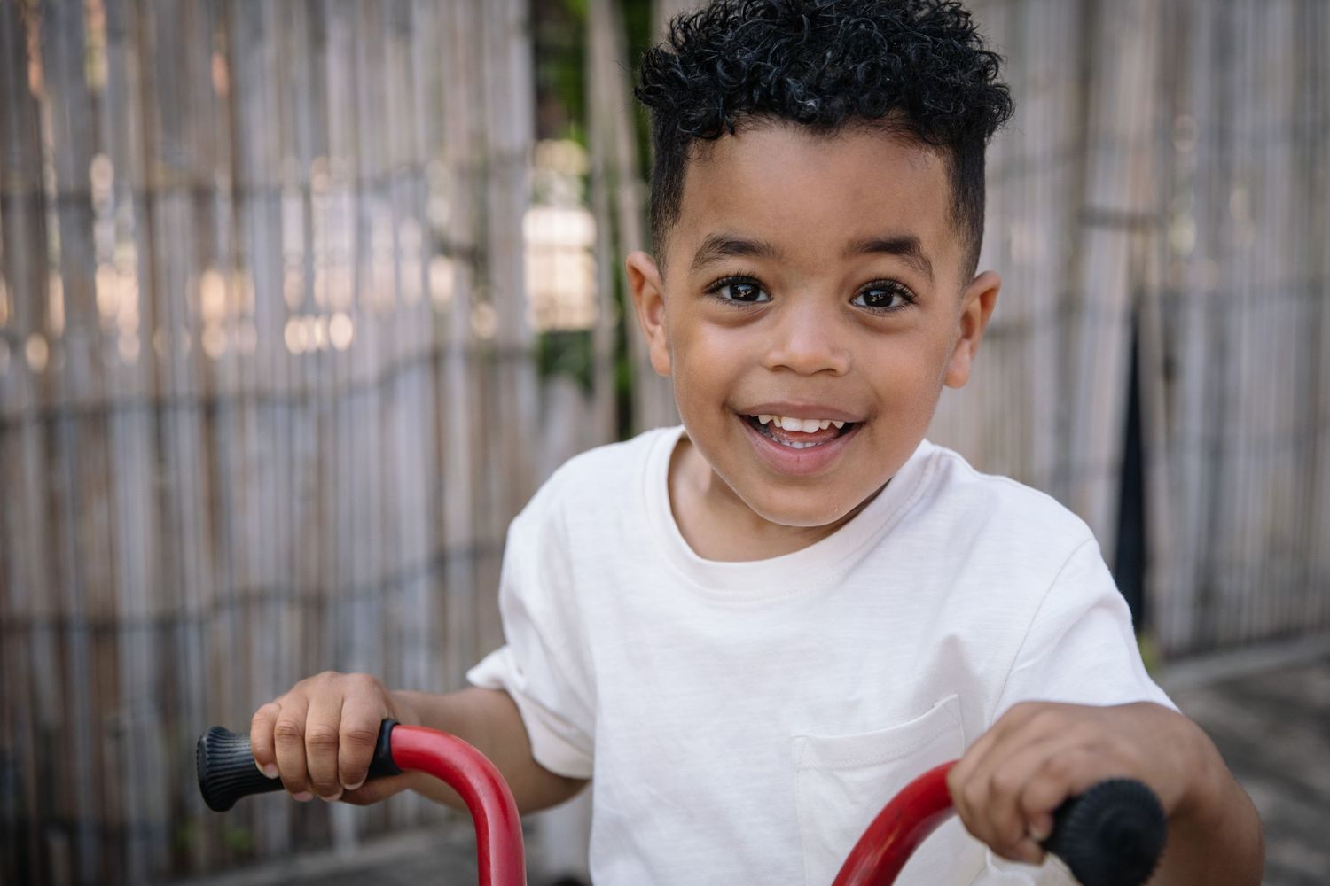 Young child in white t-shirt grips handlebars with bright smile outdoors.