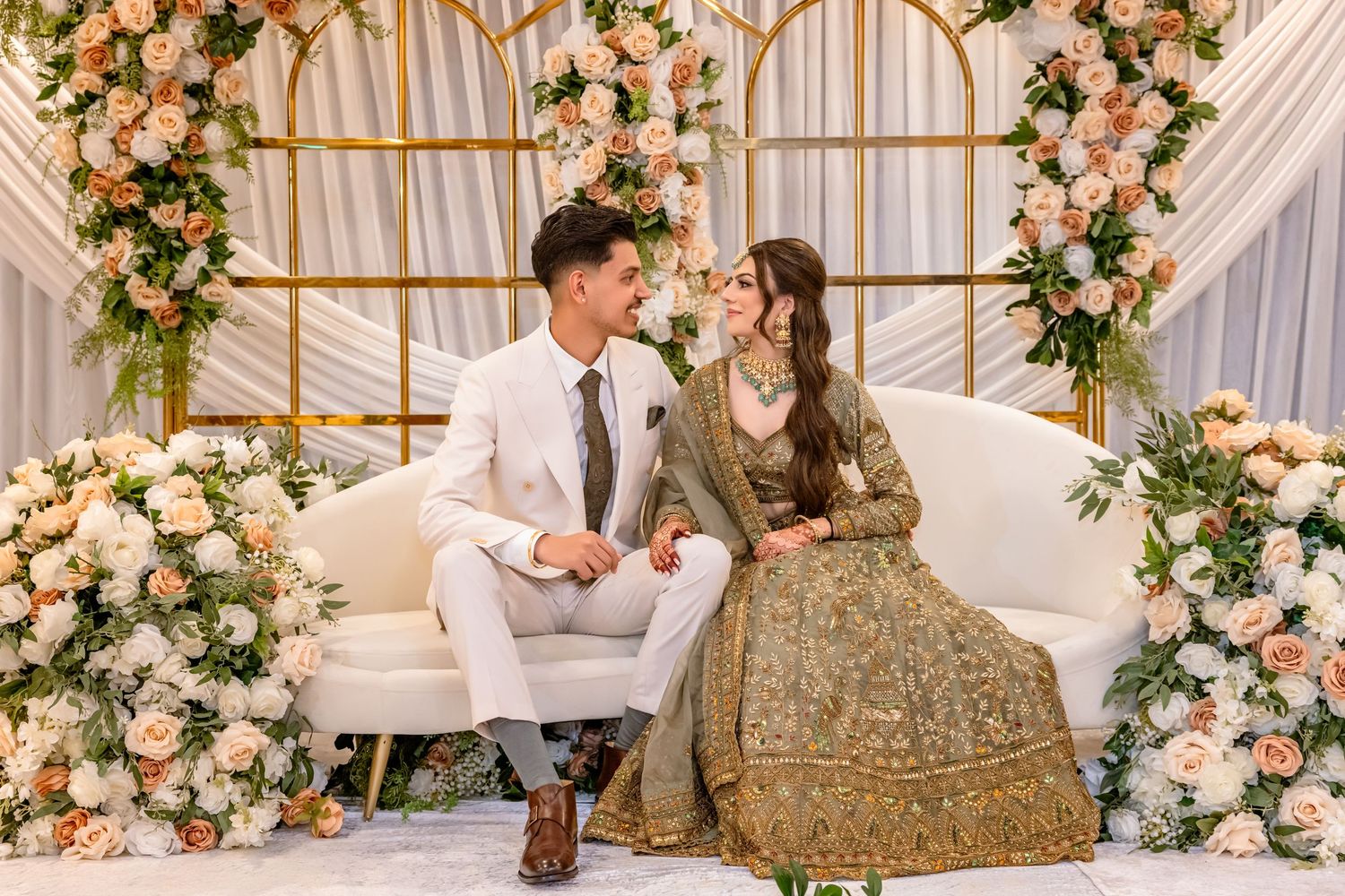 Bride and groom in elegant wedding attire seated on white sofa surrounded by peach floral arrangements.