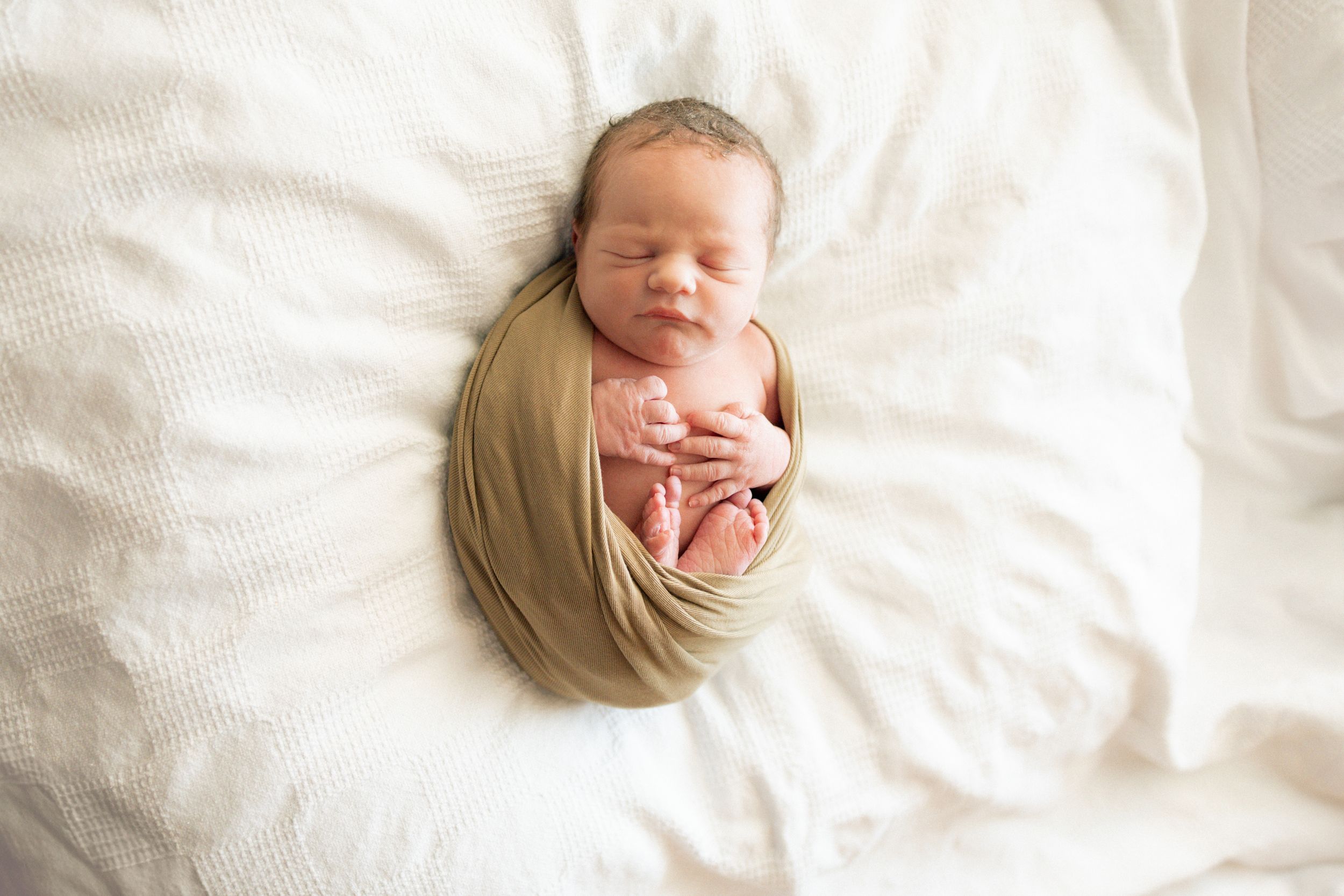 Newborn baby swaddled in tan wrap sleeping peacefully on white blanket in professional newborn photography.