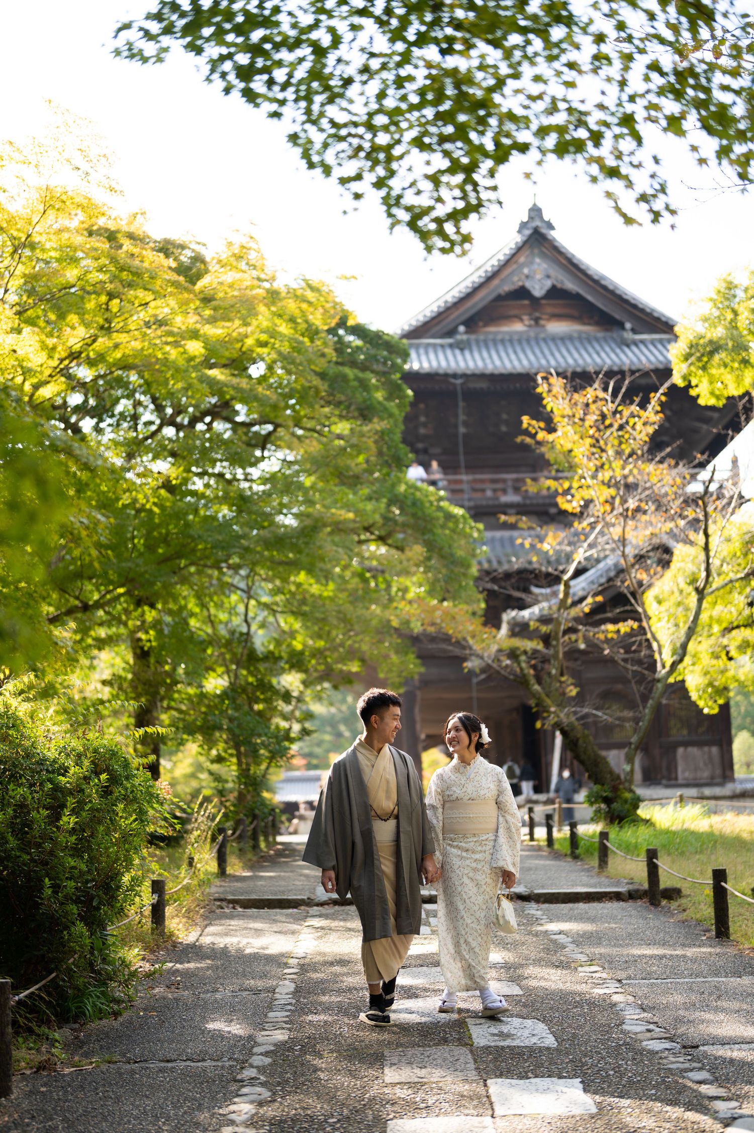 Couple in traditional Japanese kimono walking along a stone path near a historic temple surrounded by lush green trees.
