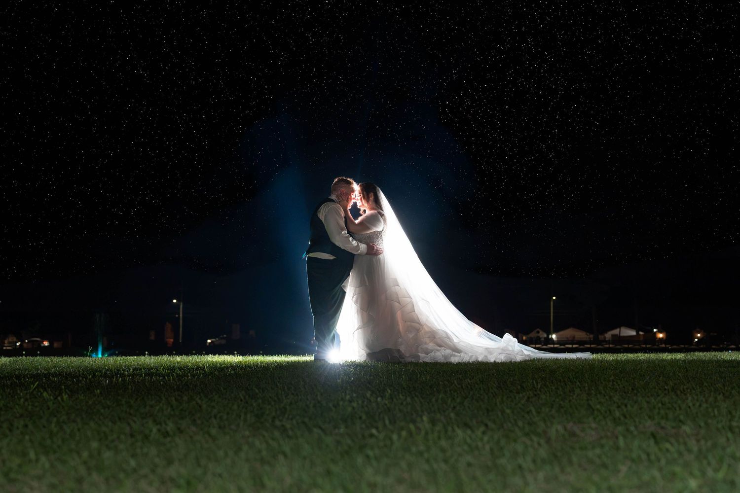 Bride and groom share an intimate embrace on a grassy field at night, backlit by a glowing light that illuminates her long wedding veil.