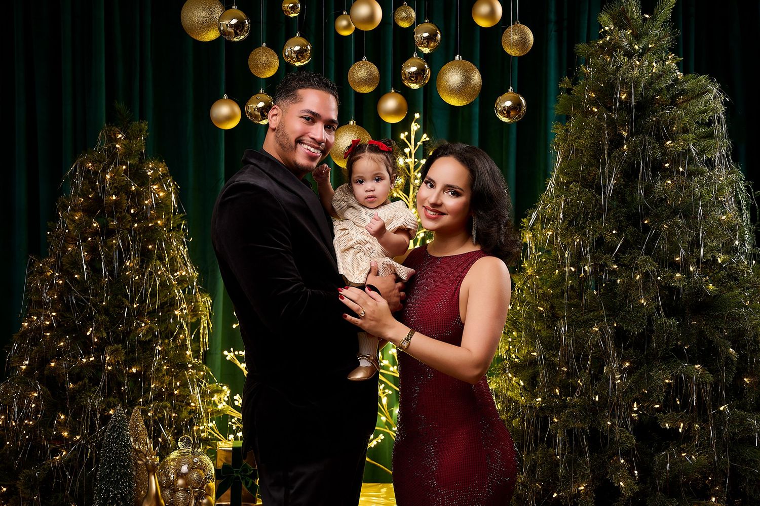 Family posing with baby amid Christmas trees and golden ornaments in festive holiday portrait setting.