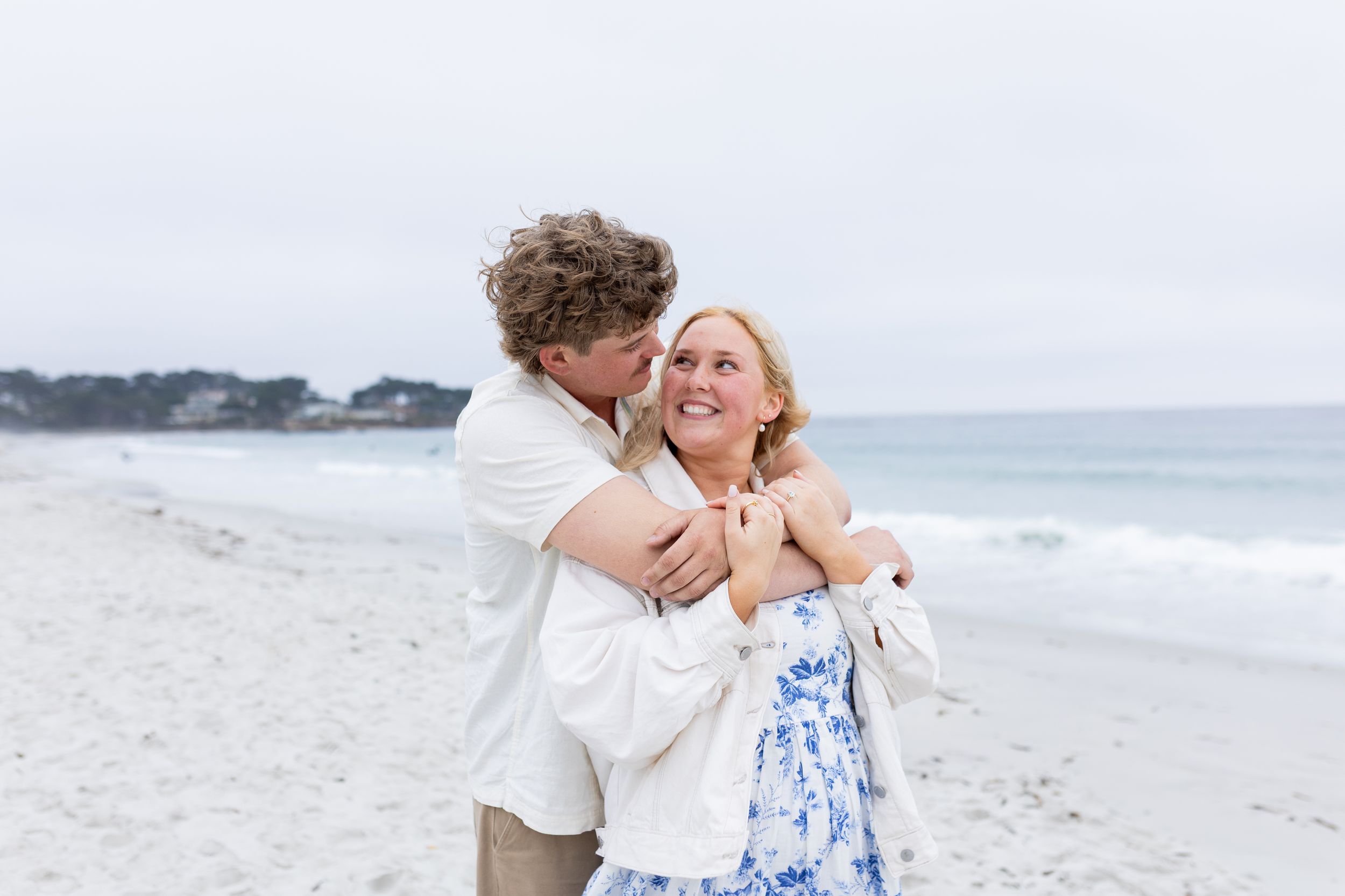 Couple embracing and laughing on a sandy beach with ocean waves, woman in blue floral dress, man in white shirt.