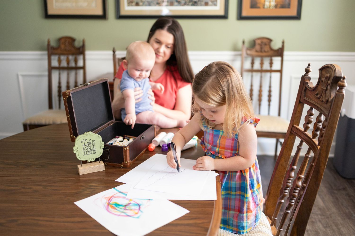 Woman holding baby watches young girl drawing with markers on white paper at wooden dining table.