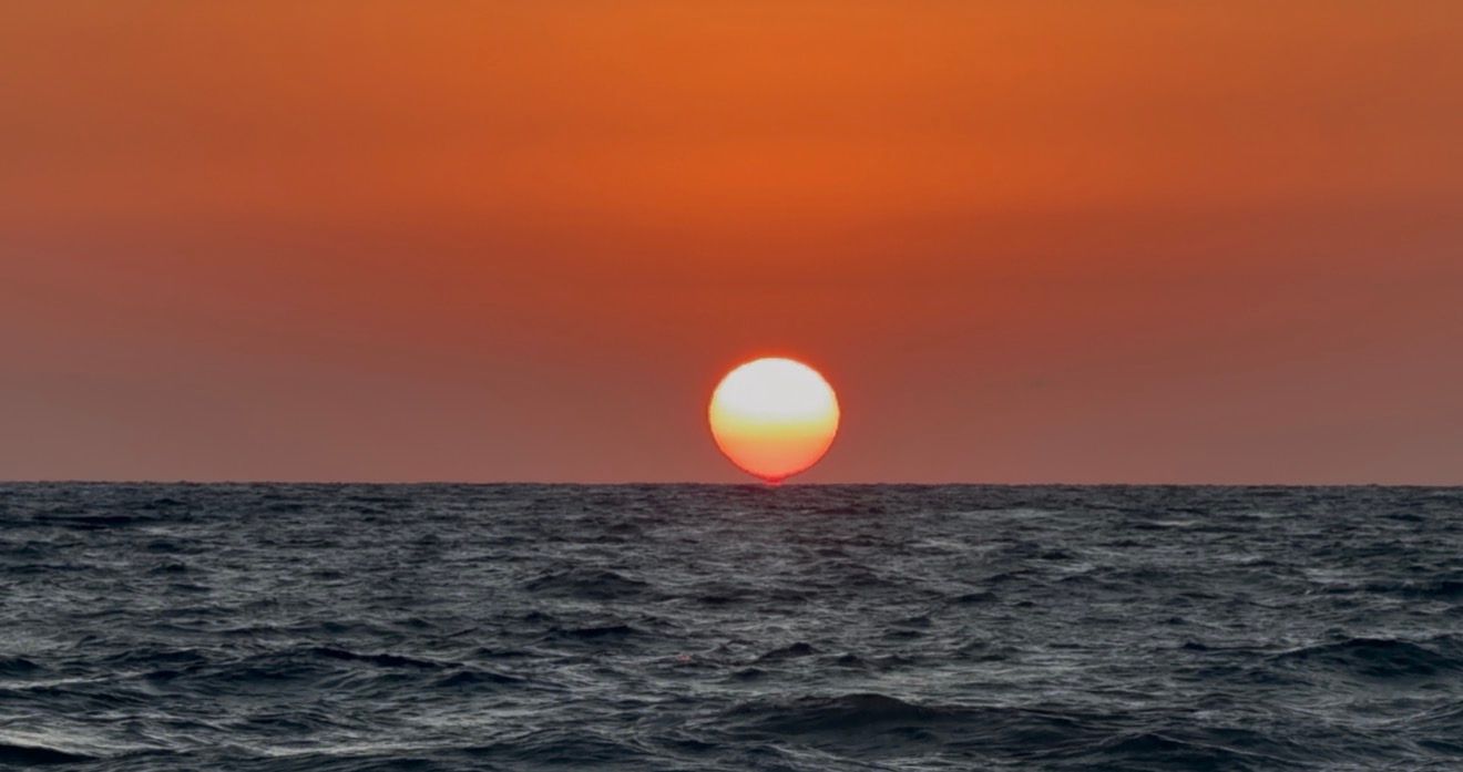 A vibrant ocean sunset with the sun touching the horizon, casting deep orange and red hues across the sky and sea.