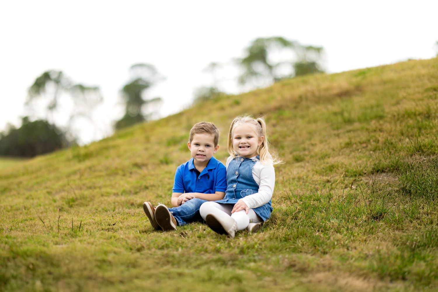 Two young children sitting together on a grassy hillside outdoors, smiling happily in casual blue outfits.