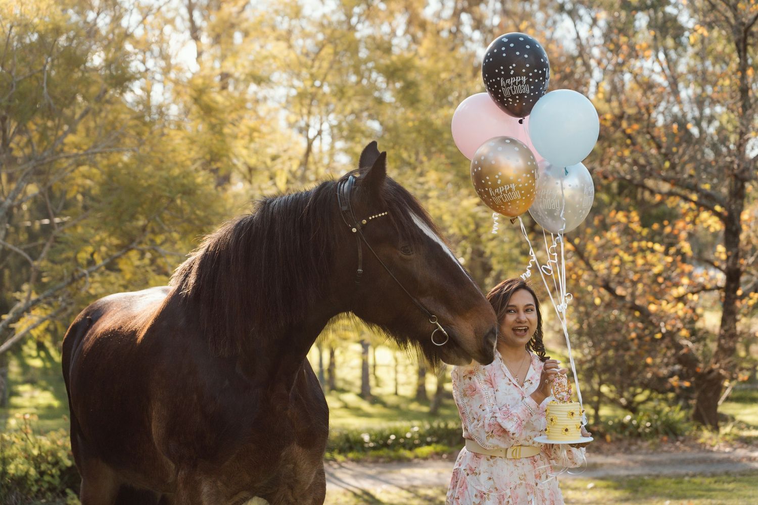Young woman holding balloons beside a brown horse in an autumn park setting during a birthday photoshoot with a horse in sydney