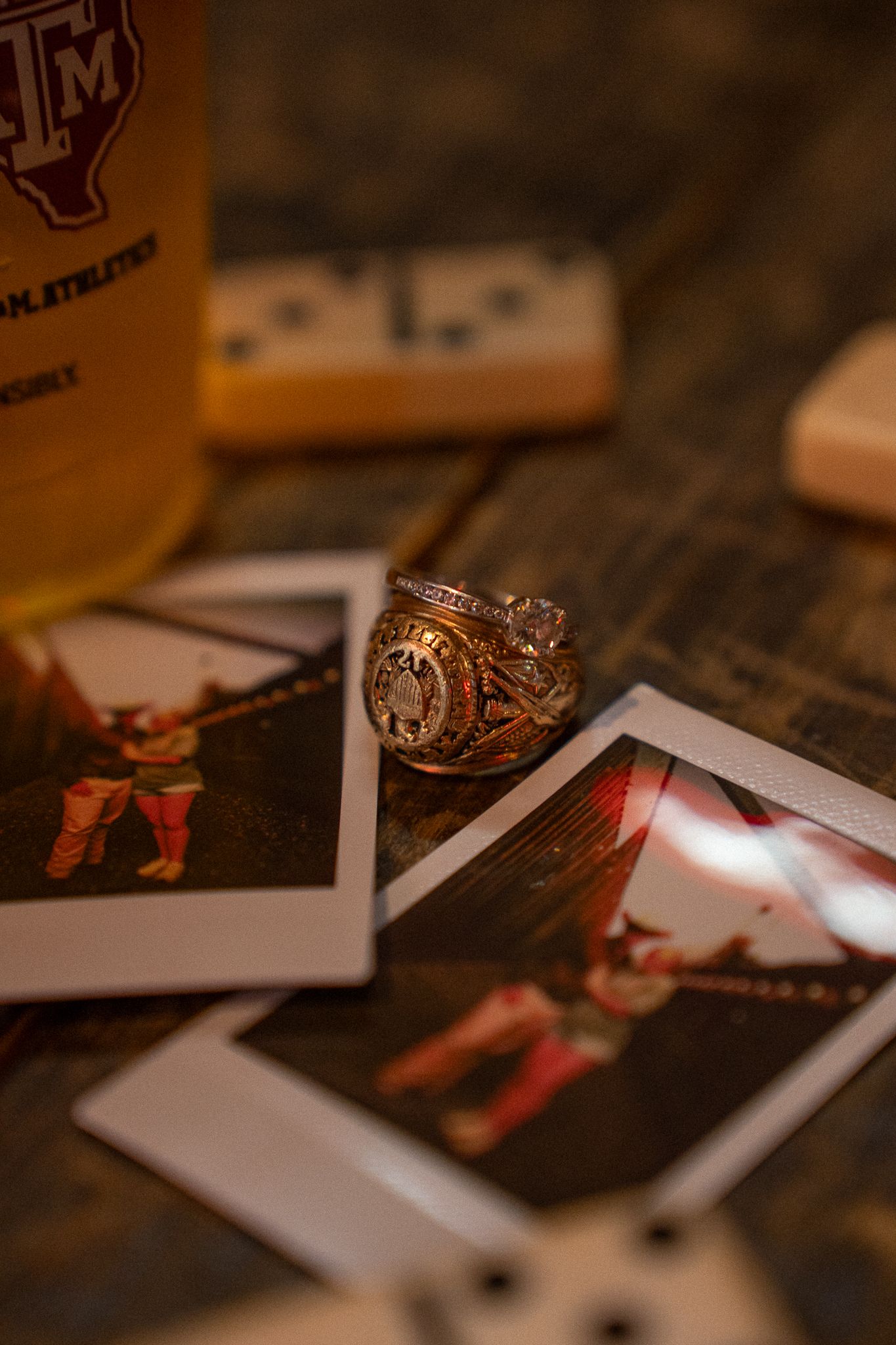 Texas A&M Aggie ring resting on Polaroid photos with a beer bottle and dominoes on a rustic wooden table.