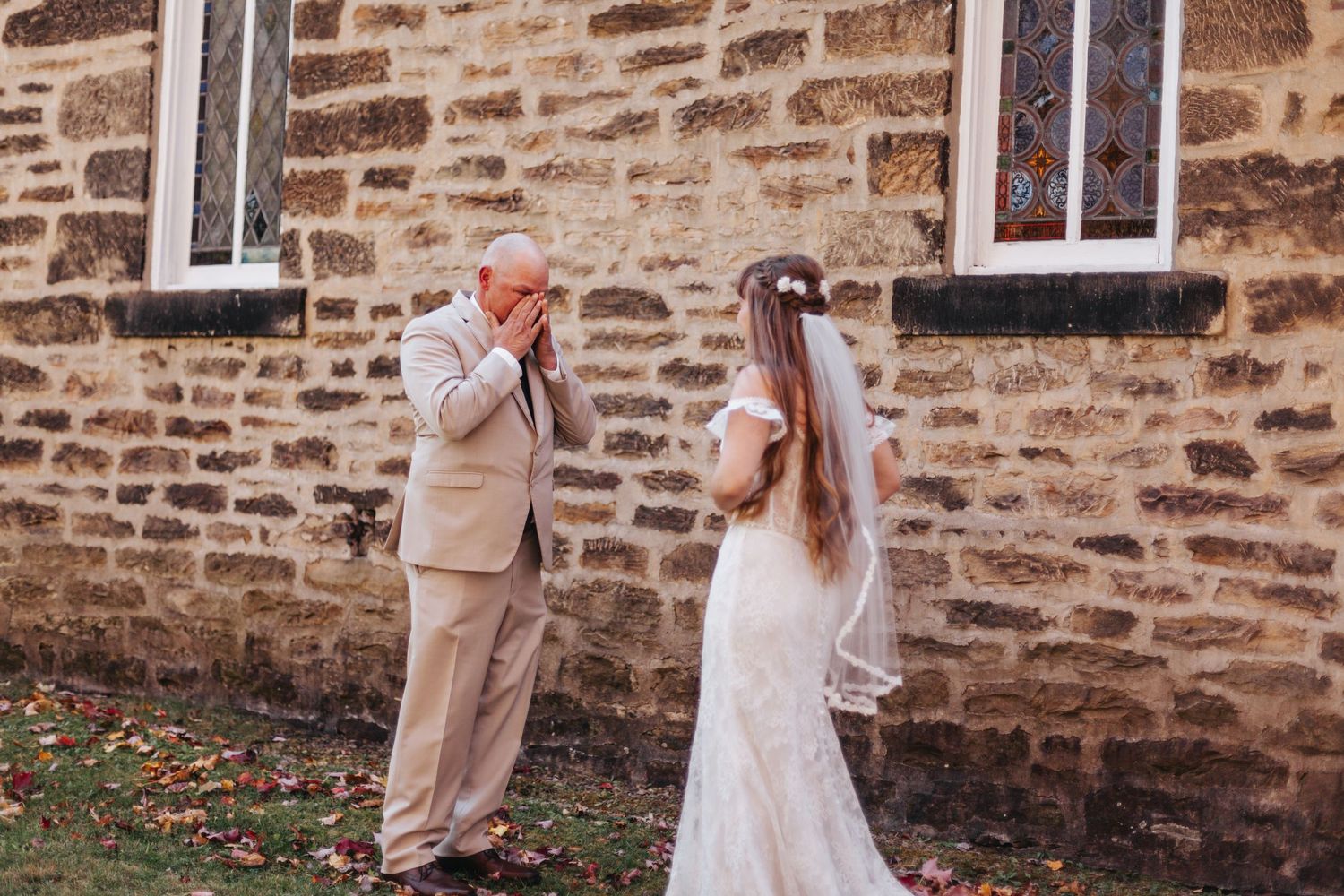 Emotional father covers his mouth seeing bride daughter in wedding dress outside stone church.