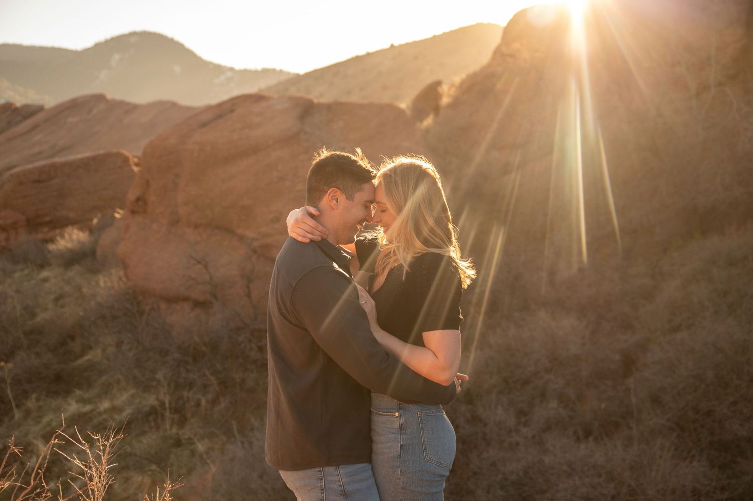 A couple embraces in a sun-drenched desert landscape during golden hour with mountains in the background.