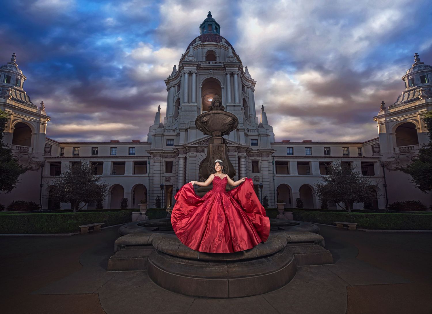 A dramatic red ball gown stands out against the ornate Pasadena City Hall architecture at dusk with stormy clouds overhead.
