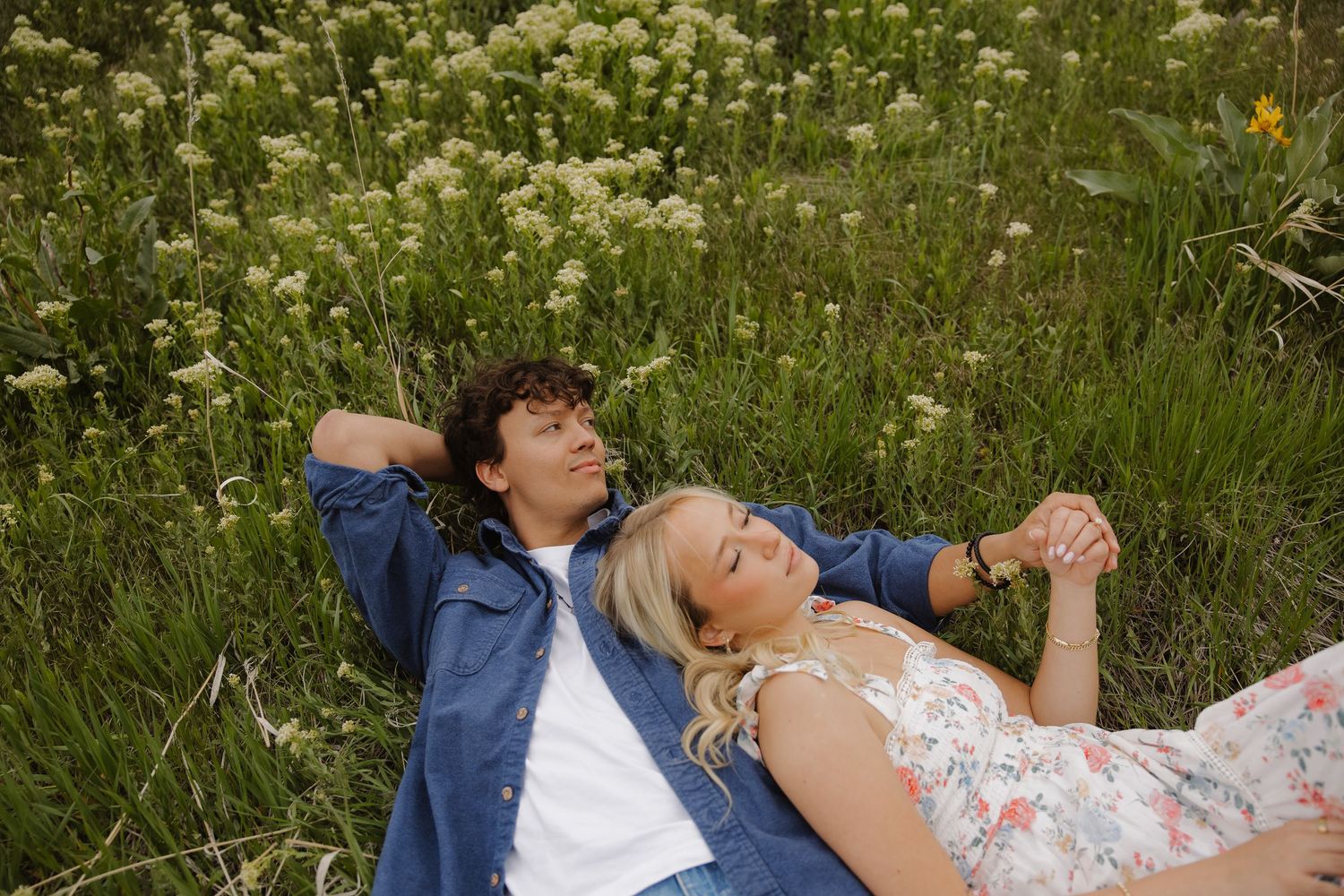 Couple lying in a lush green meadow holding hands surrounded by white wildflowers.