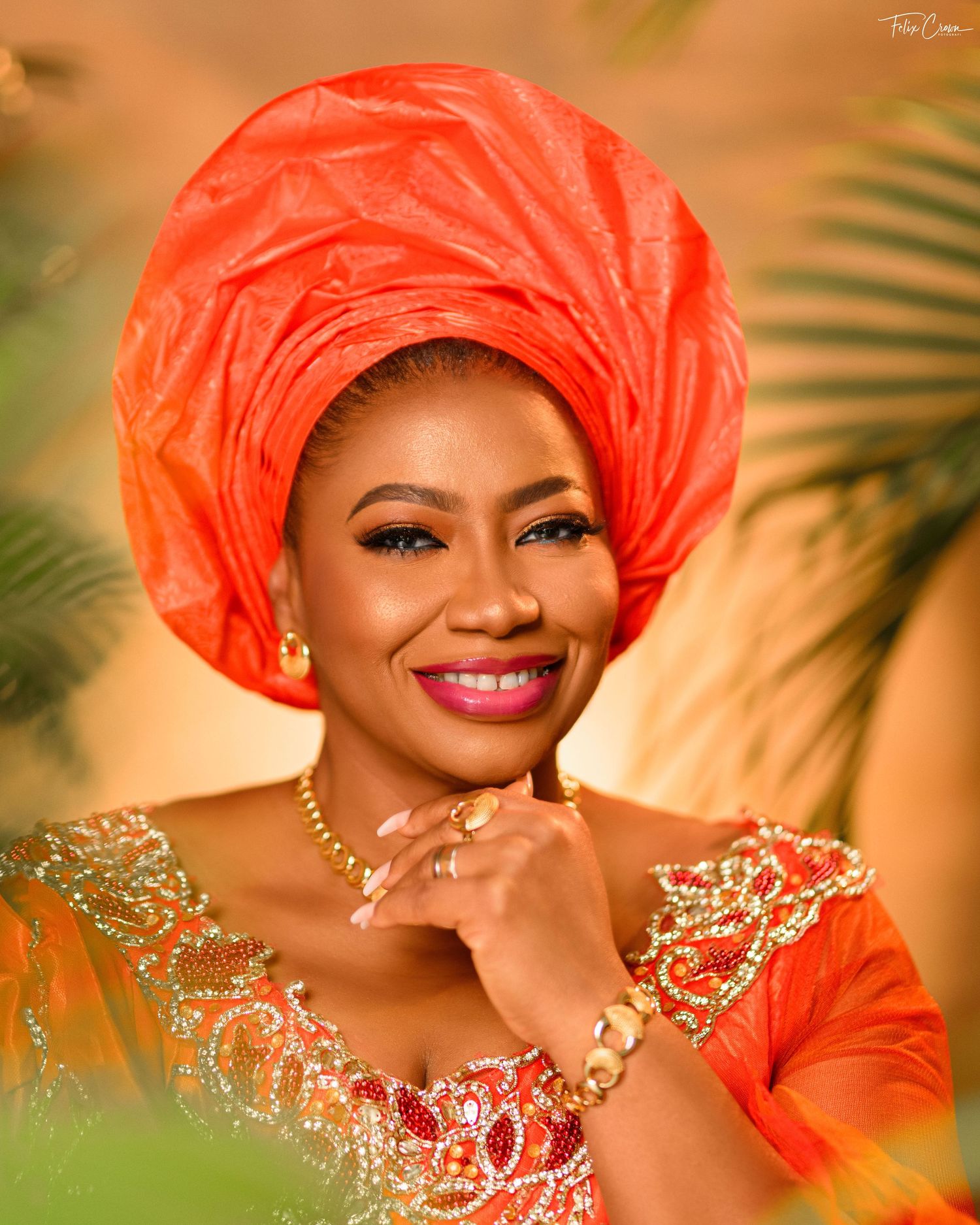 Traditional formal dress in vibrant coral with beaded details against warm golden bokeh background and palm fronds.
