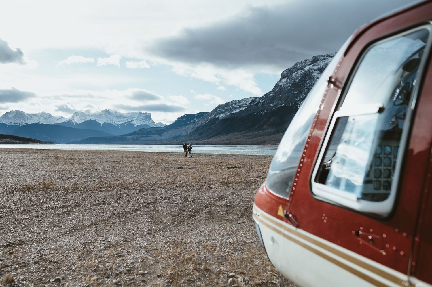 Red helicopter parked on barren landscape with snow-capped mountains in distance.