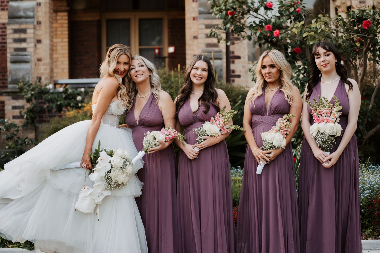 A wedding party poses together outdoors wearing purple bridesmaid dresses holding white floral bouquets.