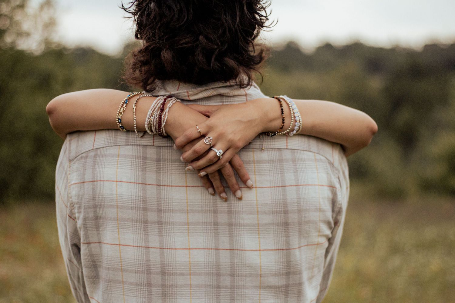 Arms with multiple rings and bracelets wrapped around a plaid shirt in a field at sunset.