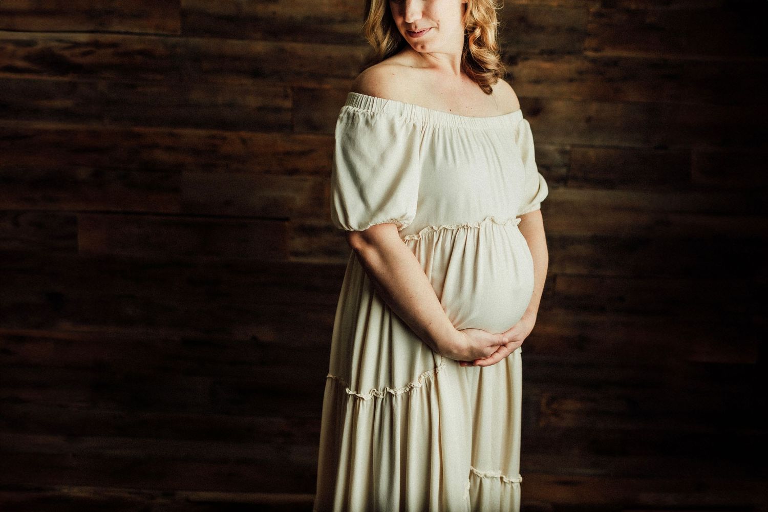 Maternity photo session in cream dress against dark wooden backdrop.