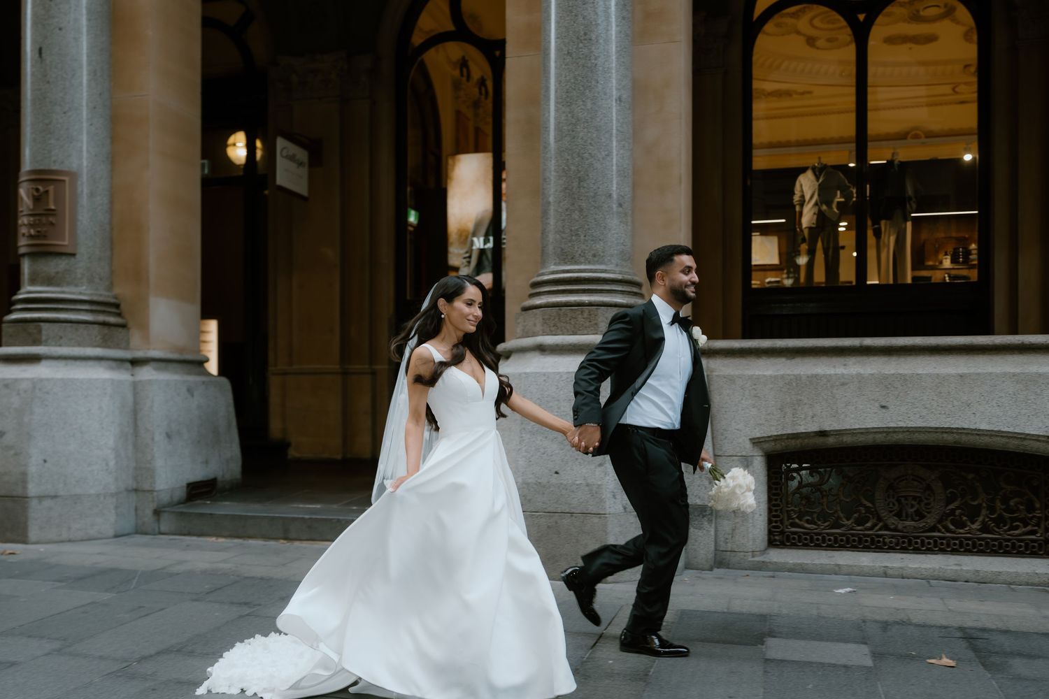 A couple in wedding attire dancing joyfully together in front of an elegant columned building entrance.