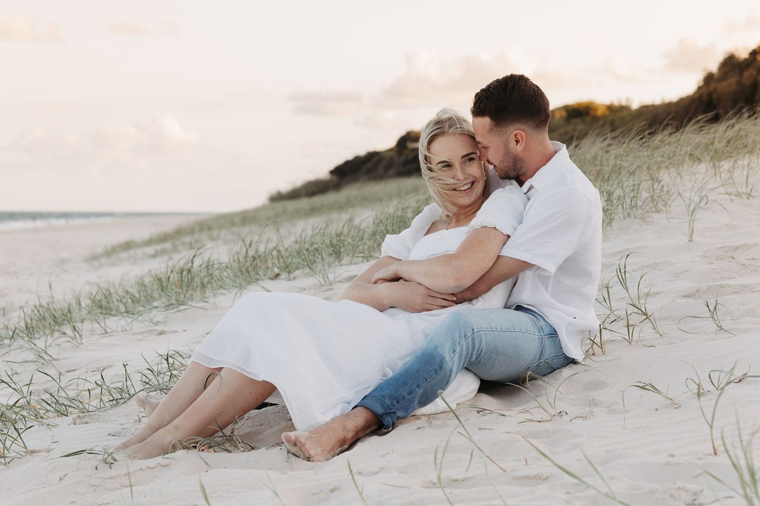 Couple in casual white clothing embraces while sitting together on sandy beach dunes at sunset.
