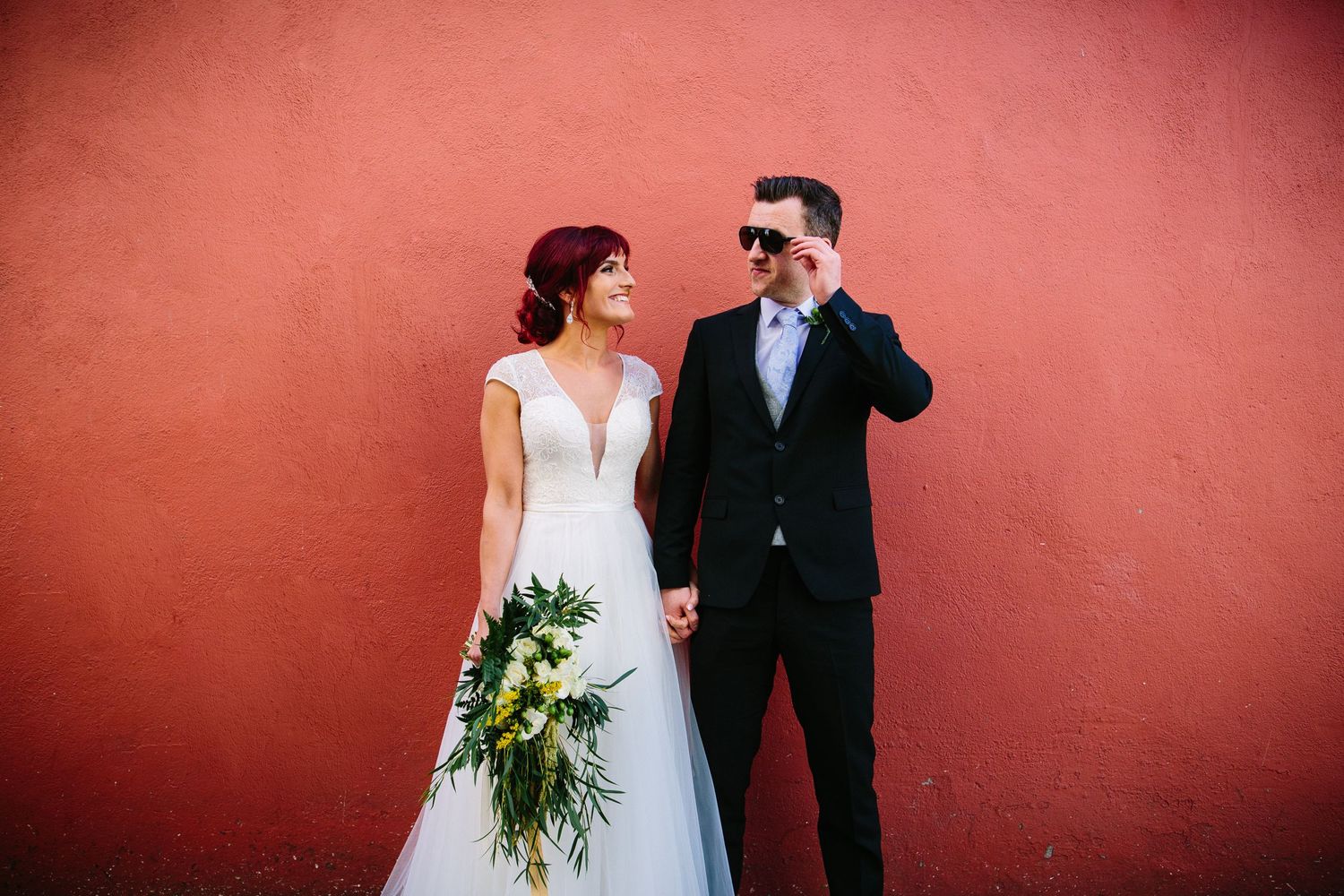 A couple in wedding attire stands against a red wall while the bride holds a cascading green and white bouquet.