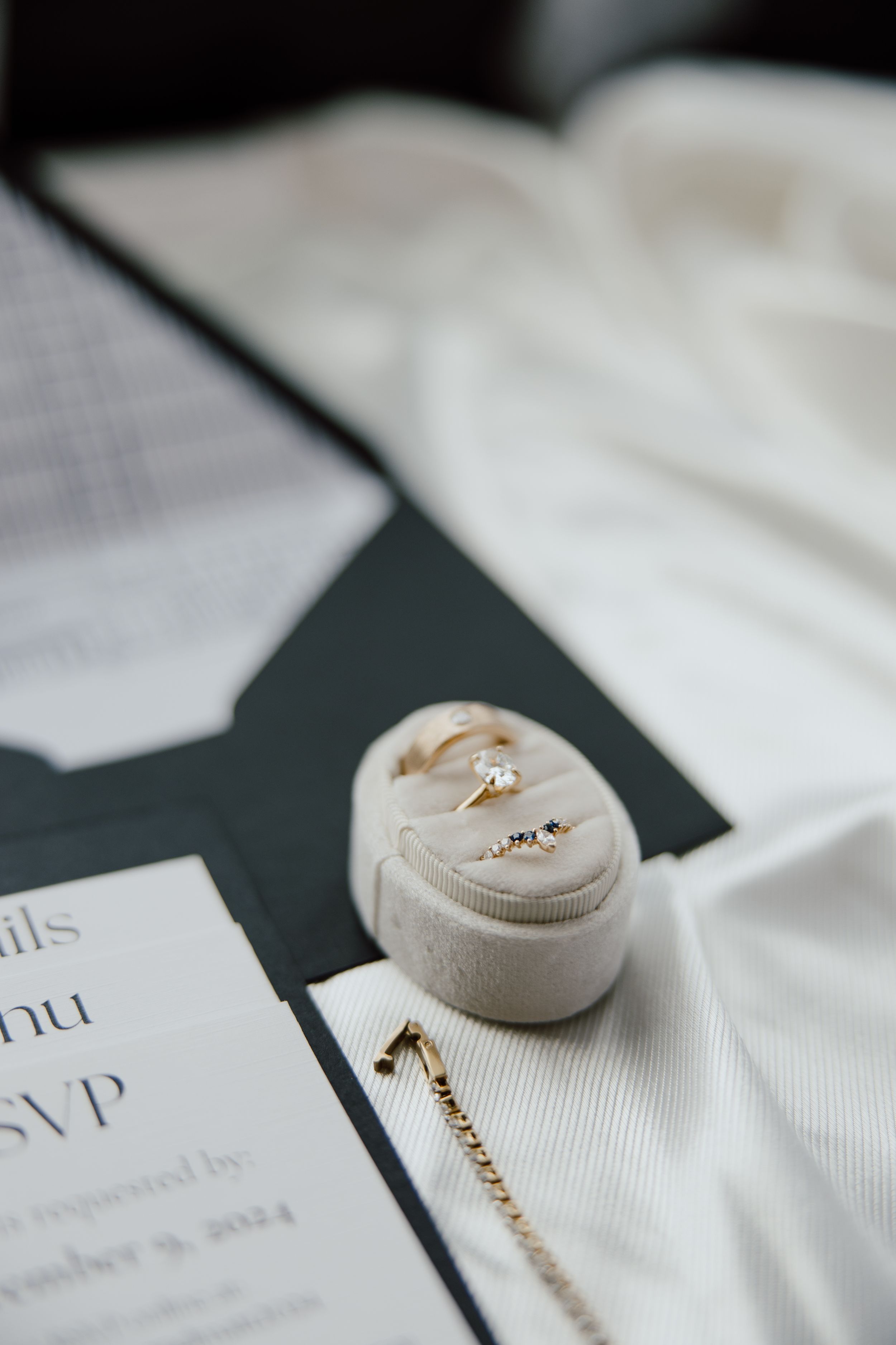 Wedding rings rest in a beige velvet ring box next to invitation cards and white fabric.