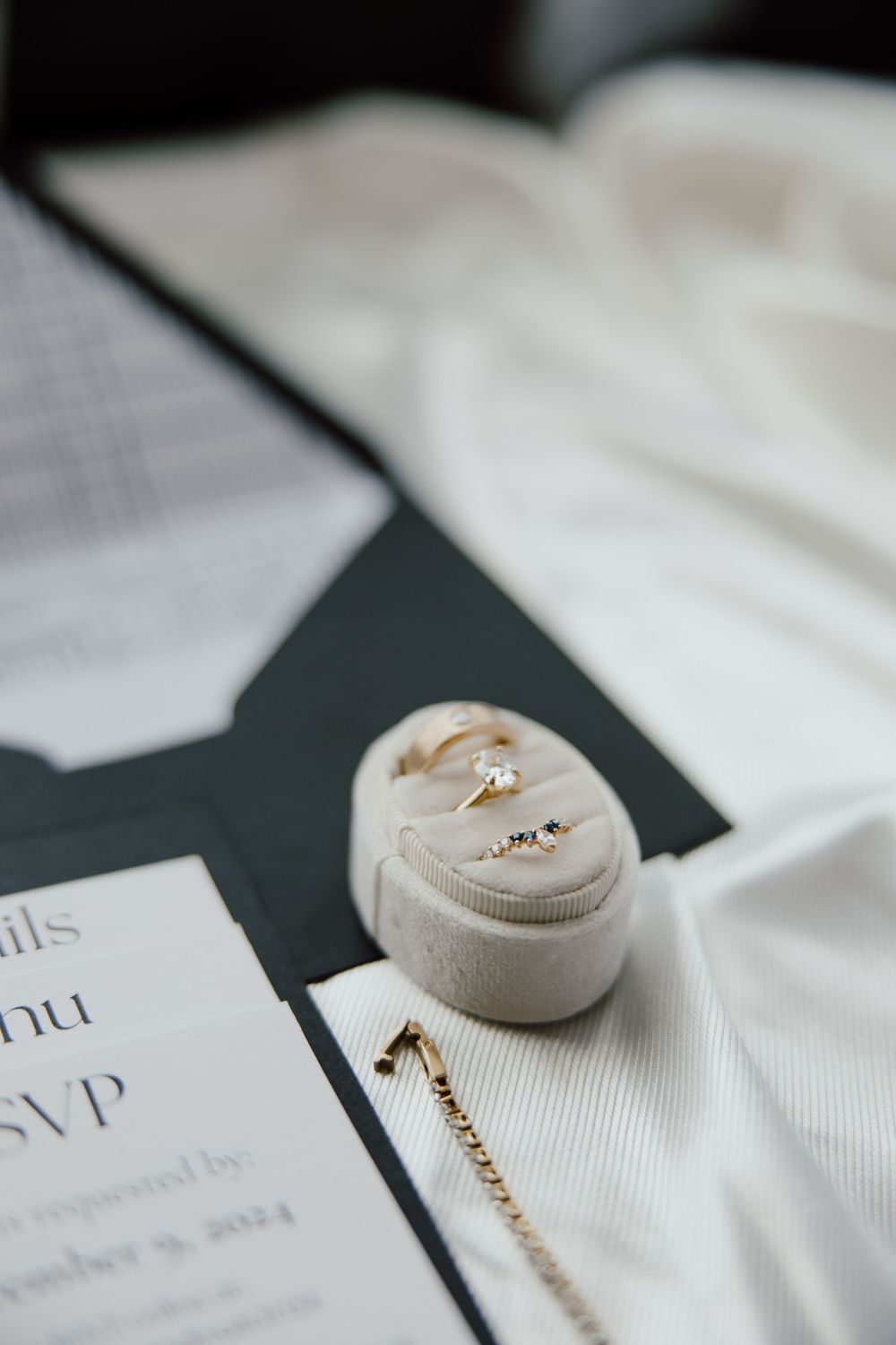 Wedding rings rest in a beige velvet ring box next to invitation cards and white fabric.