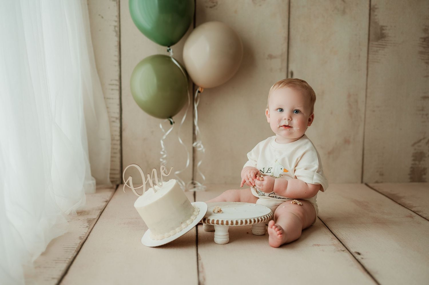A small child sits on a white wooden floor next to a white cake and green and white balloons for a birthday celebration.