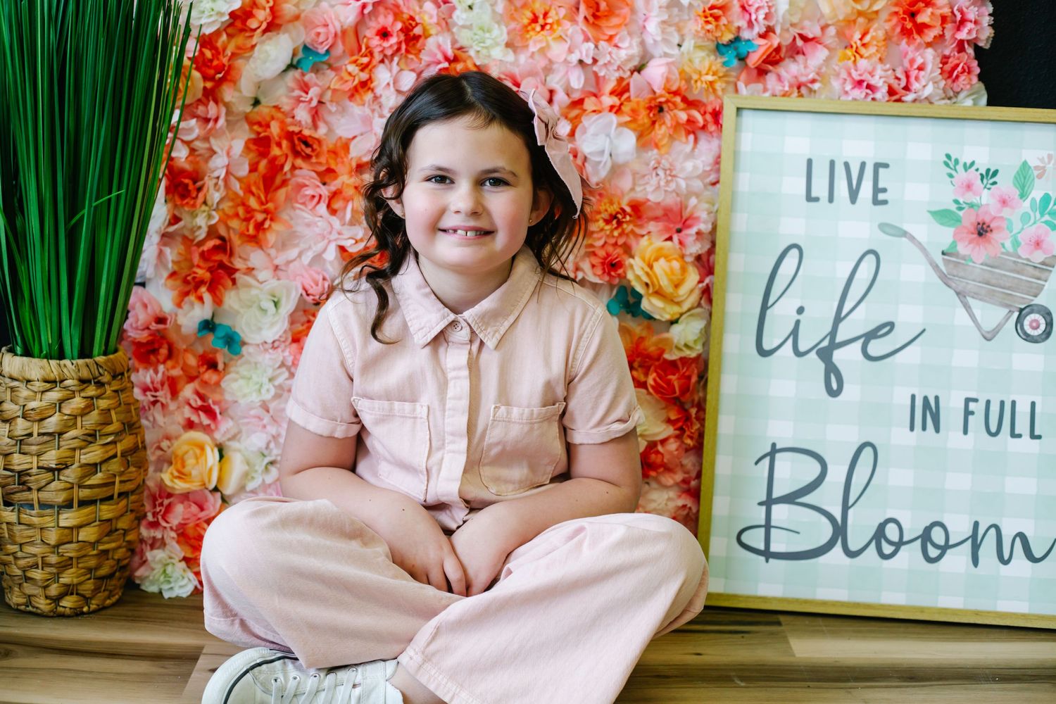 A seated figure in pink clothing poses against a coral floral backdrop with a 'Live Life in Full Bloom' sign and potted plant.