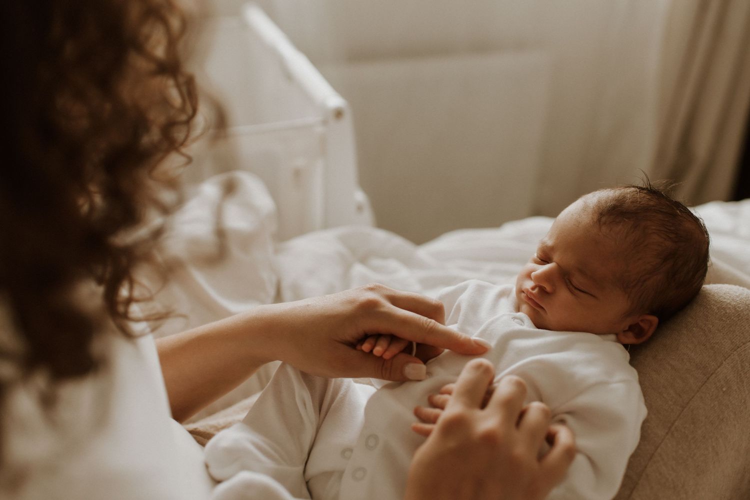A gentle hand reaches out to comfort a peaceful newborn baby resting on soft beige bedding.