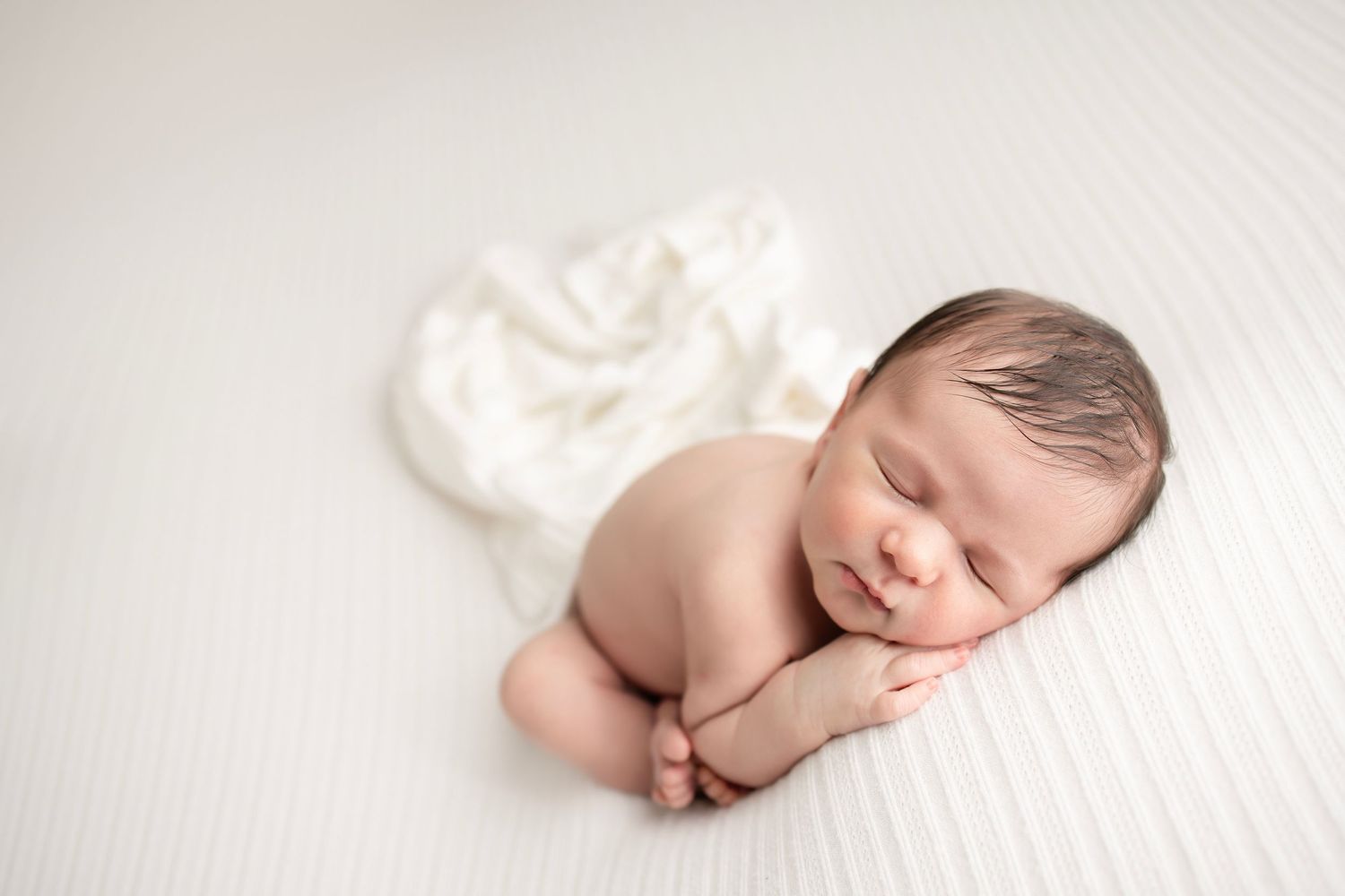 Sleeping newborn baby curled up on white textured blanket.