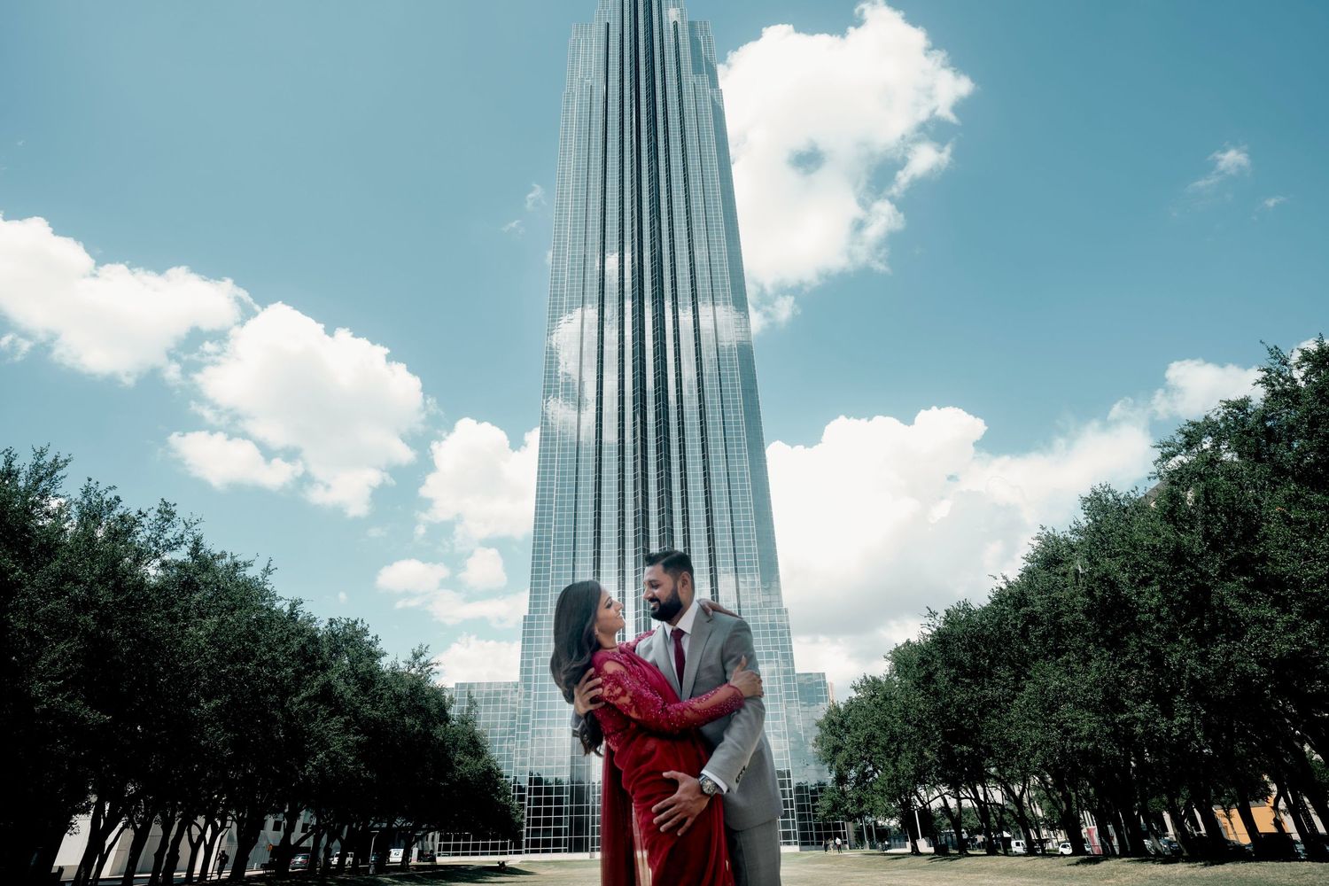 A couple poses together in front of a modern skyscraper on a bright sunny day.