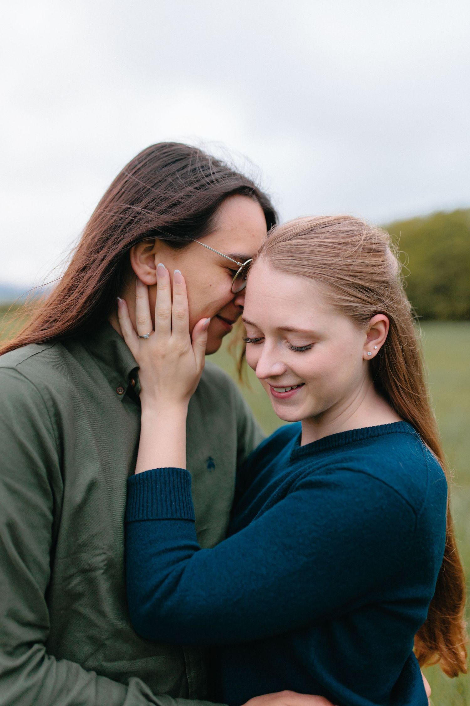 Two people share an intimate moment outdoors, embracing while wearing casual sweaters against a soft natural backdrop.