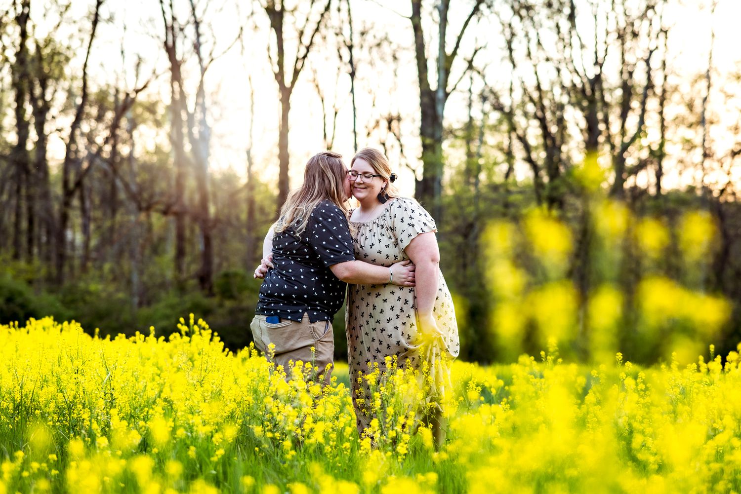 Two people share a tender embrace while standing in a vibrant yellow field of wildflowers at sunset.