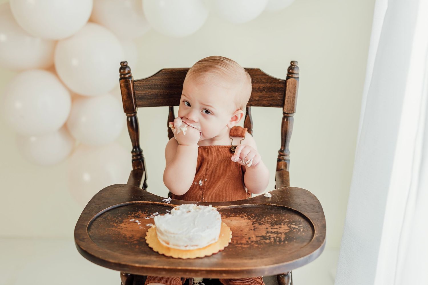 A wooden high chair with a small white cake and white balloon backdrop.