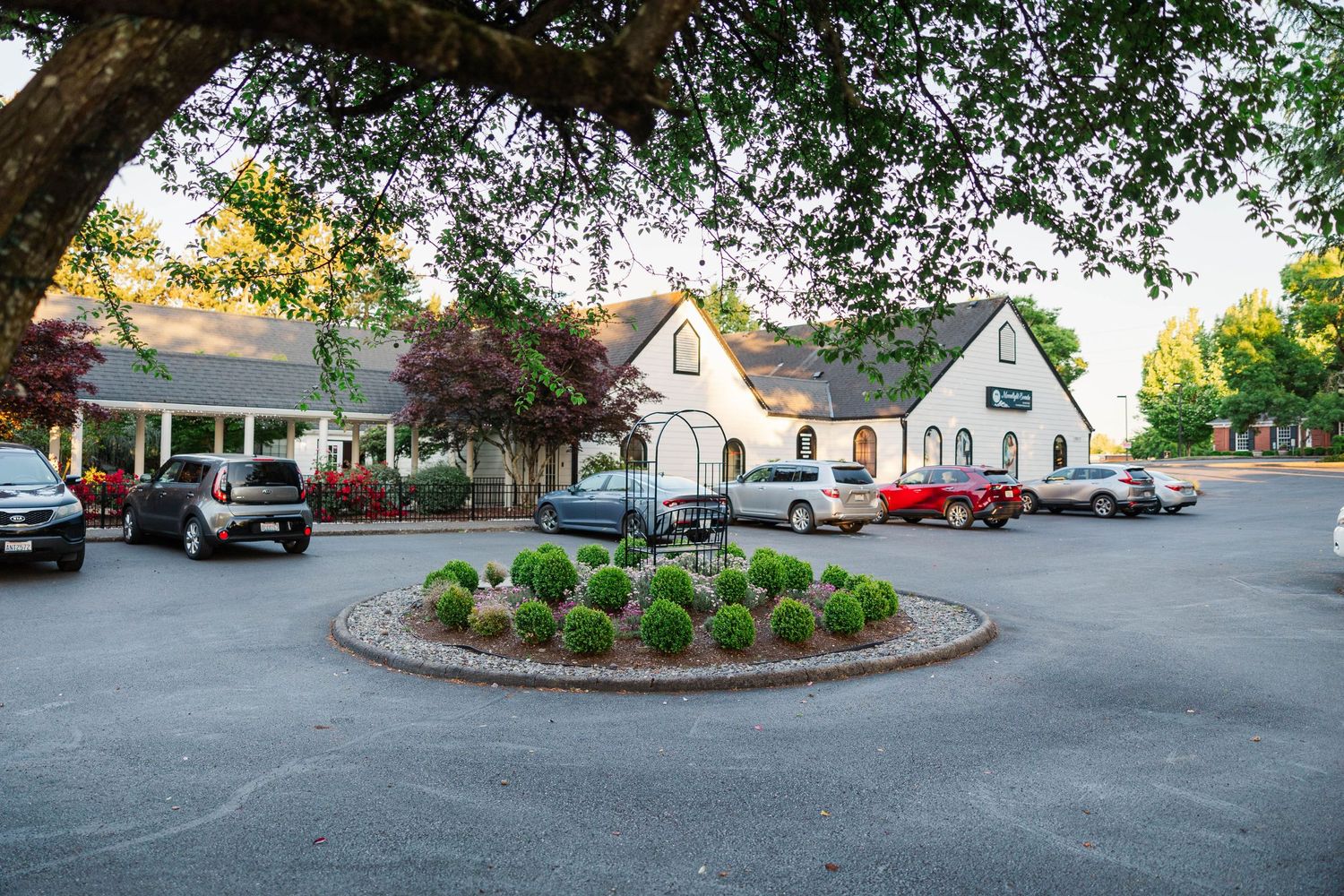 A charming white cottage-style building with a circular driveway surrounded by lush trees and landscaping.