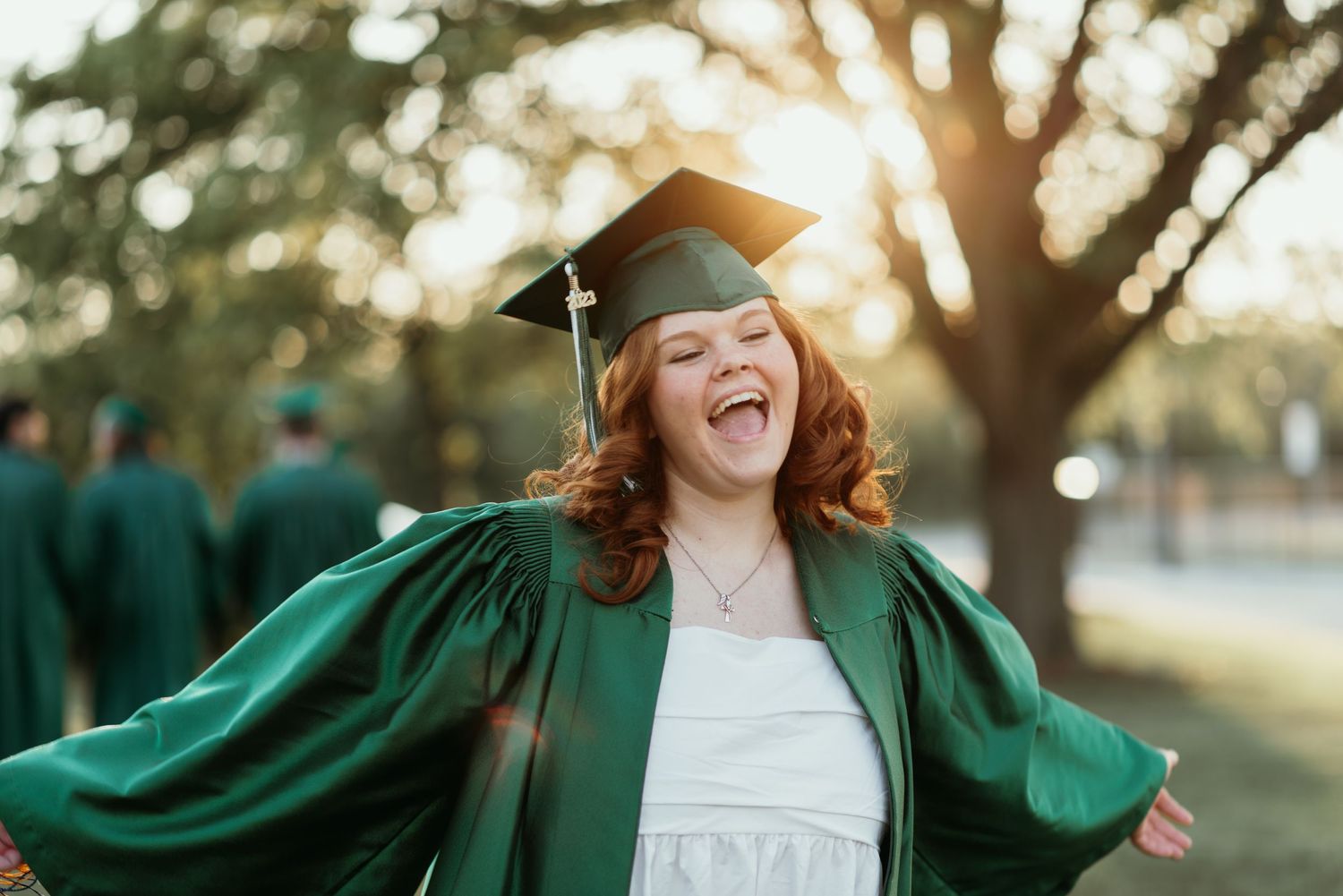 Joyful graduate in green cap and gown celebrating graduation outdoors with golden sunlight.