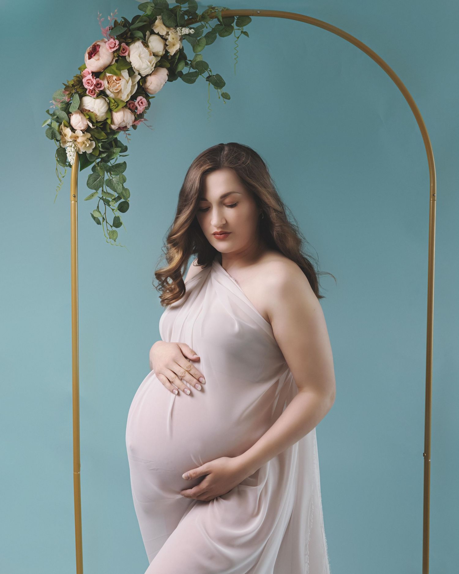 A maternity portrait with a pink gown against a teal backdrop, featuring a golden arch decorated with pink roses.