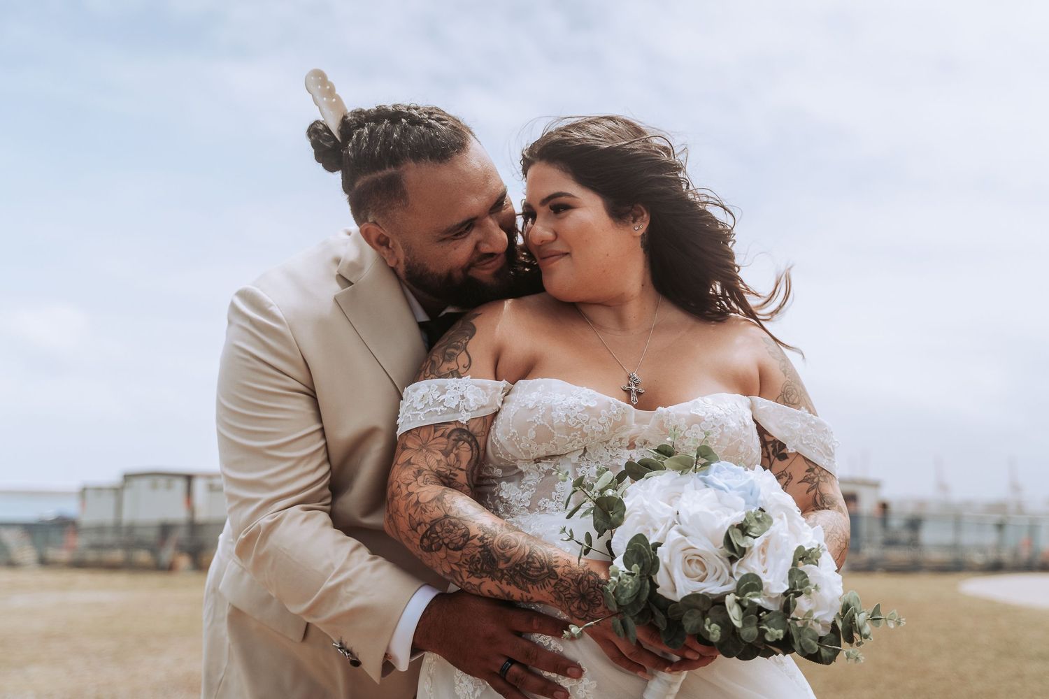 Romantic newlyweds share an intimate moment outdoors at La Perouse Sydney with white floral bouquet against cloudy sky.