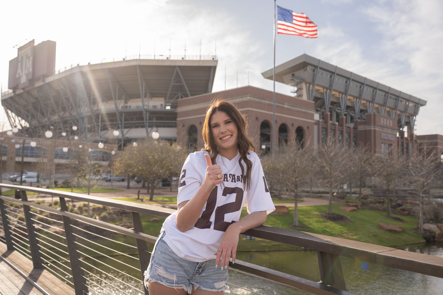 College sports fan poses on sunny spring day outside stadium with American flag flying in background.