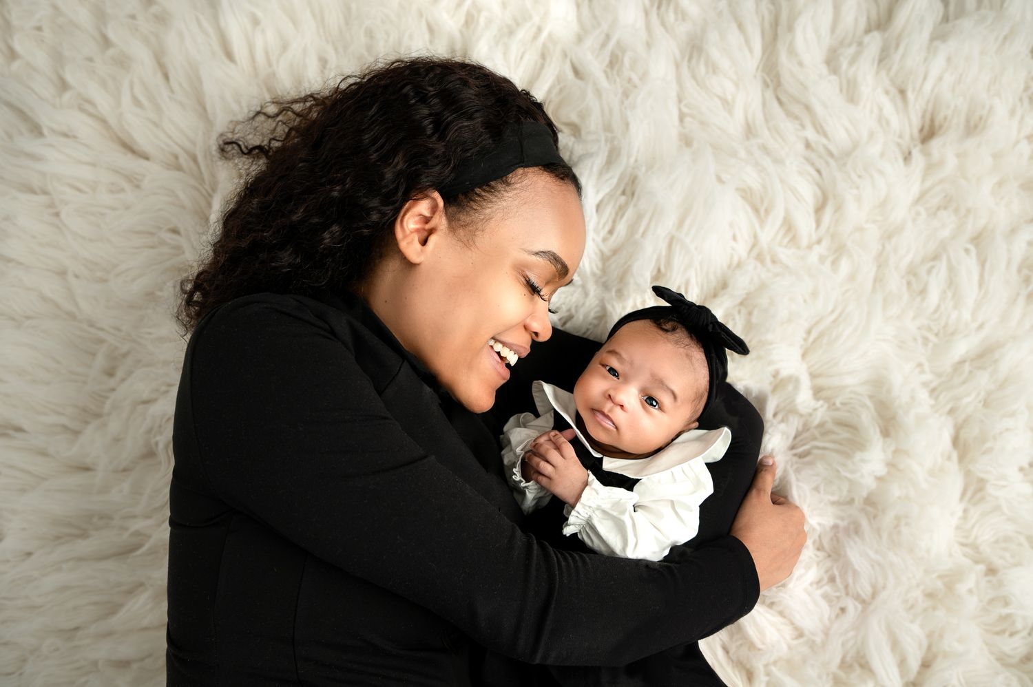 Mom and baby girl share a tender moment lying on a soft white fur backdrop during a professional photo session with Flora Levin Photography.