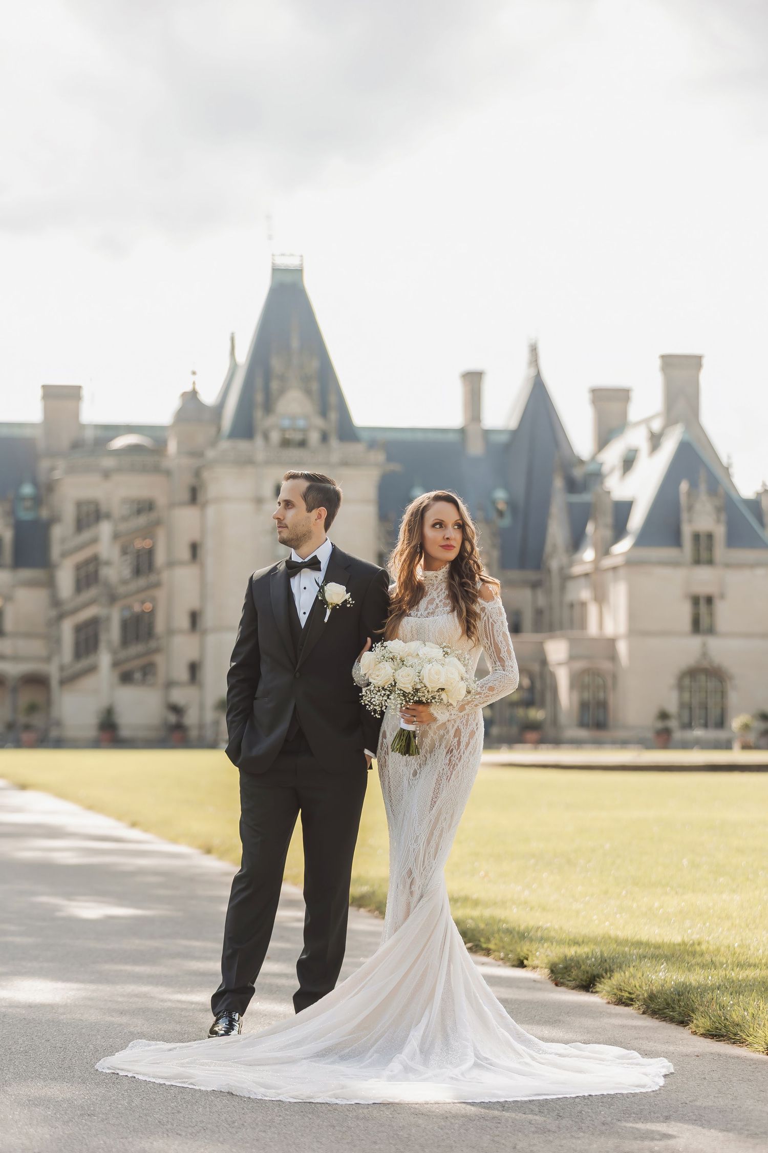 Elegant wedding couple poses in front of majestic castle with dramatic cloudy sky background.