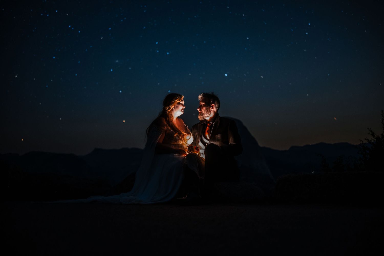 Silhouetted couple sits under starry night sky in mountains sharing intimate moment illuminated by warm light.