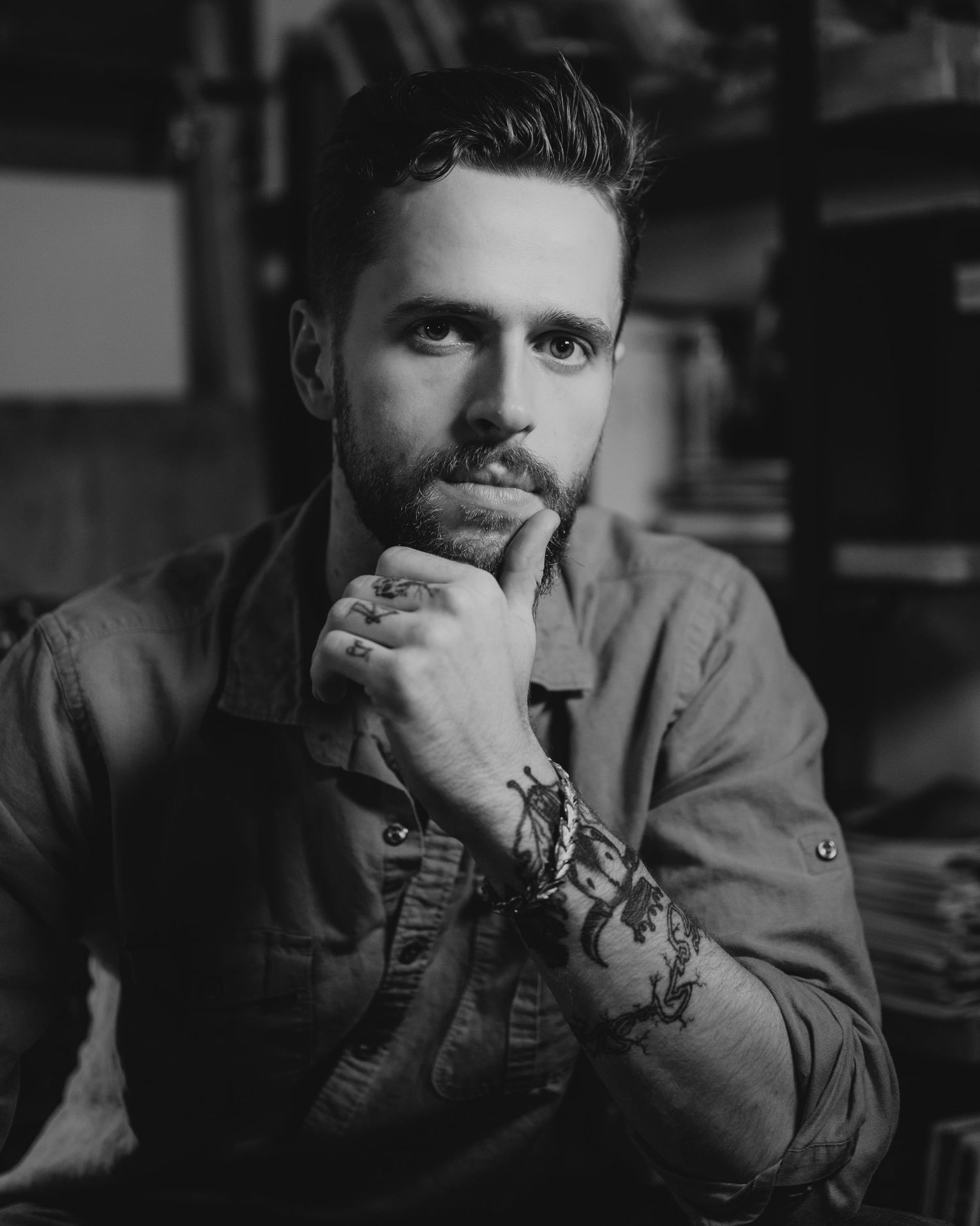 Black and white portrait of someone in casual attire with facial hair resting their chin on hand in contemplative pose.